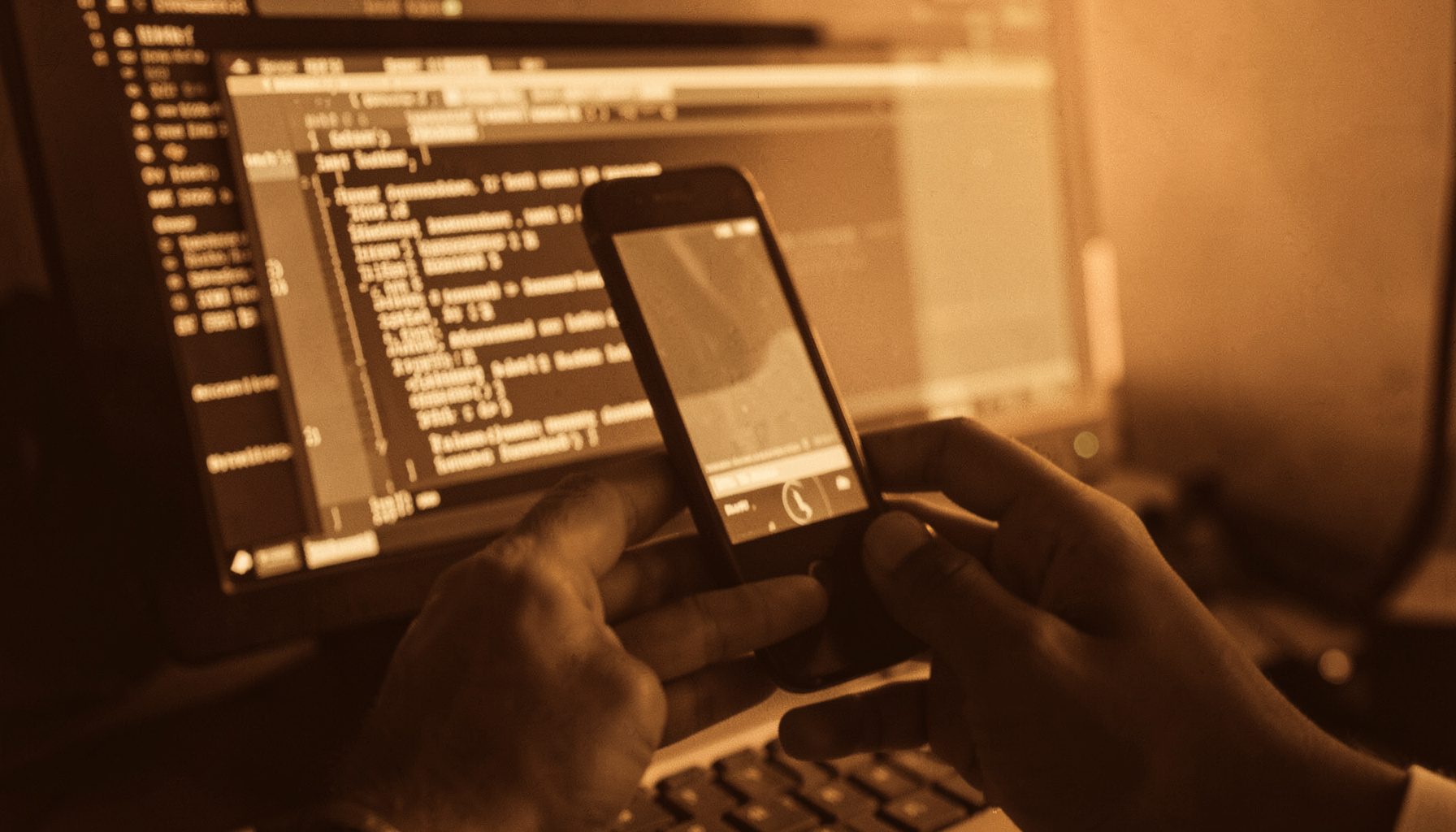 Photographie en duotone brun et crème d'une main tenant un téléphone devant un écran d'ordinateur affichant du code, tons chauds et ambiance de veille technologique